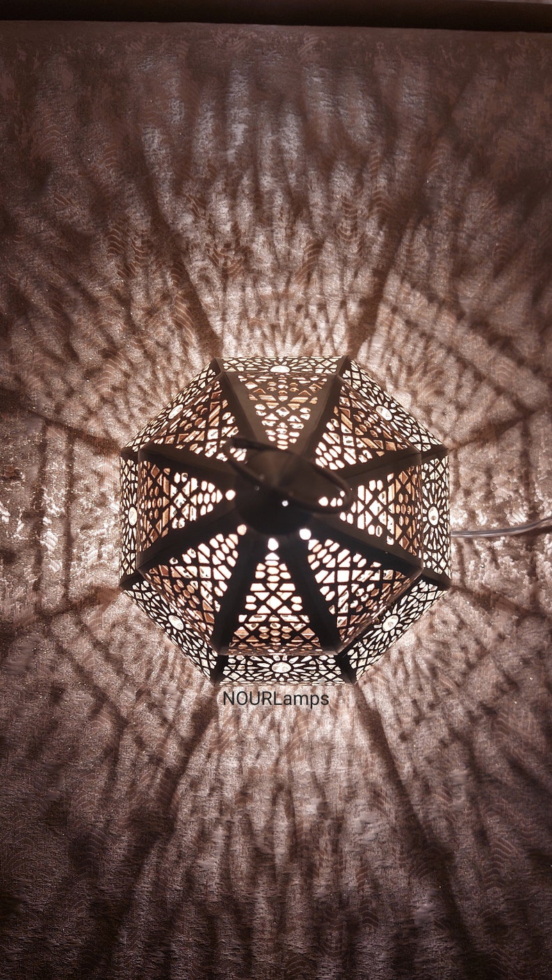 Puede incluir: Una linterna marroqu&iacute; de metal blanco con intrincados patrones geom&eacute;tricos. La linterna est&aacute; encendida y proyecta sombras en la pared detr&aacute;s de ella. El texto "NOURLamps" es visible en la parte inferior de la imagen.