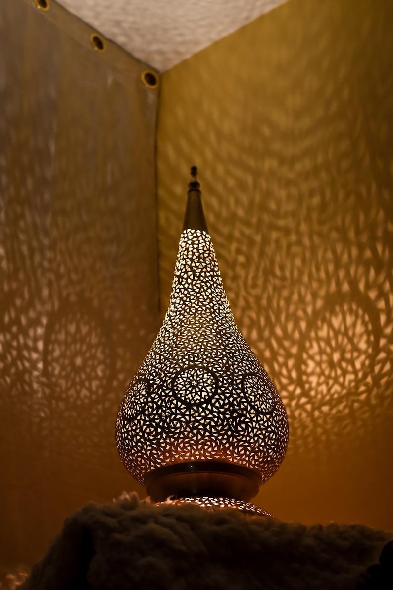 K&ouml;nnte beinhalten: Eine tropfenf&ouml;rmige Metalllampe mit einem komplizierten, lasergeschnittenen geometrischen Muster. Die Lampe strahlt ein warmes, goldenes Licht aus und wirft detaillierte, komplexe Schatten auf die umliegenden Fl&auml;chen.