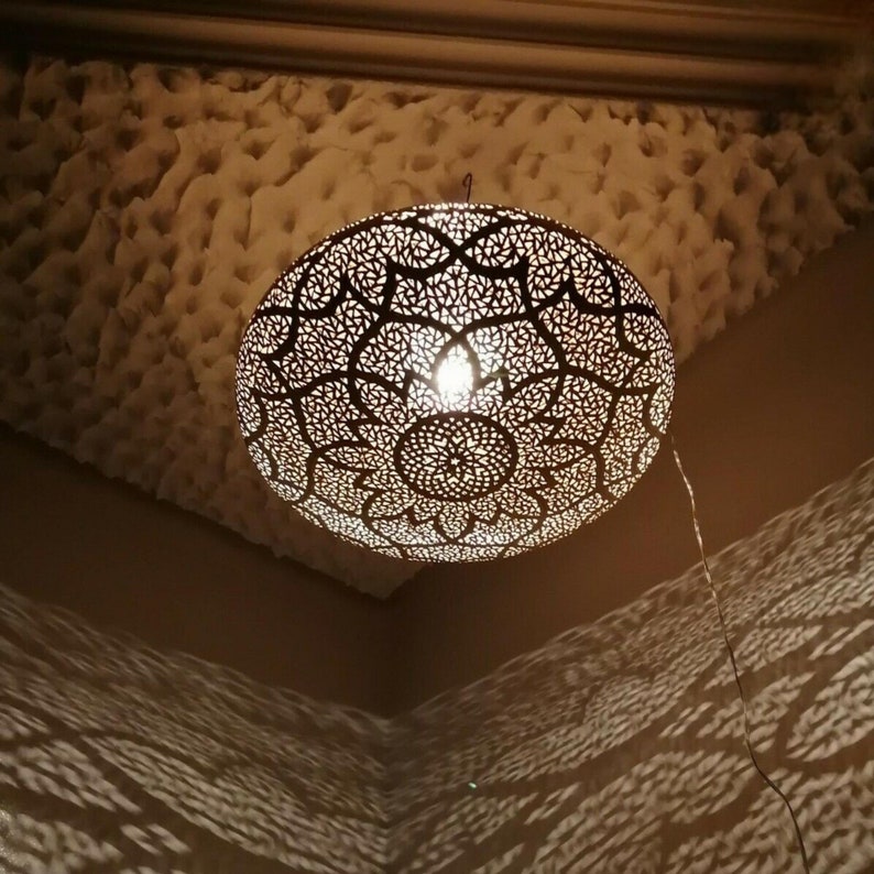 May include: A decorative, spherical pendant light with intricate cut-out patterns, casting patterned shadows on the wall. The light fixture is suspended from the ceiling, illuminating the room with a warm glow. The ceiling has a textured surface.