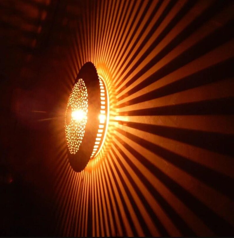 Op de afbeelding: Een ronde, decoratieve lamp met een metalen frame en ingewikkelde uitsnijdingen. Het licht werpt een warme, oranje gloed door de uitsnijdingen, waardoor een patroon van strepen op de muur erachter ontstaat.