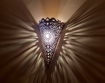 May include: A cone-shaped wall sconce featuring an intricate laser-cut pattern that emits a bright light. The metal structure casts long, dramatic, radiating shadows across the surrounding area, creating a distinct starburst effect from the light.