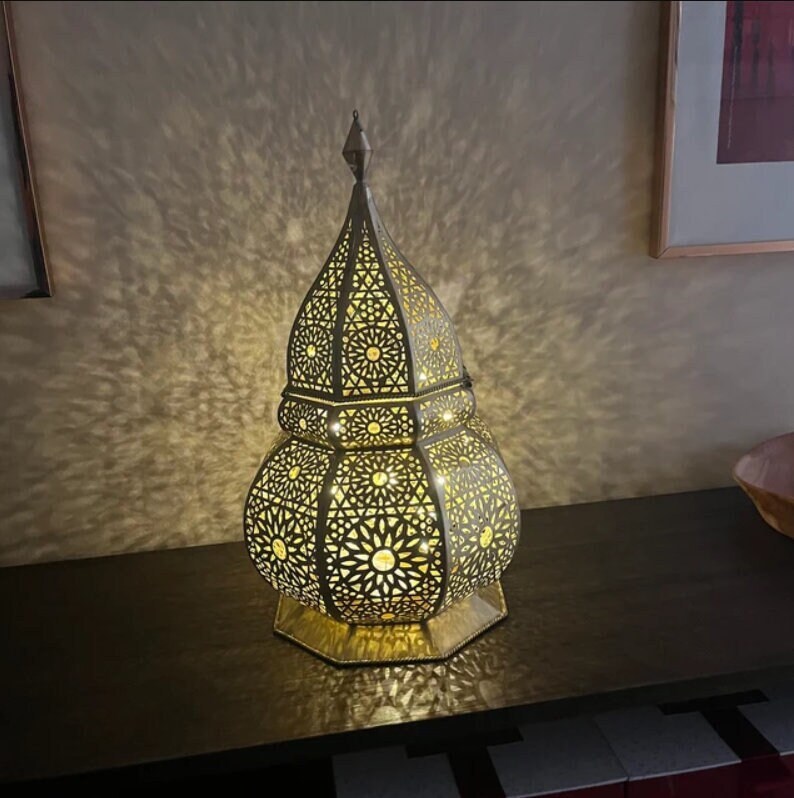 Puede incluir: Una l&aacute;mpara de metal de color dorado, alta y adornada, con un dise&ntilde;o geom&eacute;trico. La l&aacute;mpara est&aacute; encendida y proyecta un brillo c&aacute;lido.