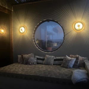 Op de afbeelding: Een knusse woonkamer met een pluche bruine bank en drie ronde wandlampen met een zonnestralenontwerp. De lampen zijn verlicht en werpen een warme gloed in de kamer. Een groot rond raam met uitzicht op de buitenwereld is zichtbaar op de achtergrond.