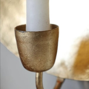 May include: A gold metal candle holder with a white candle. The candle holder has a textured surface and a curved hook.