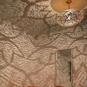 May include: An ornate, gold-colored pendant light fixture with intricate cut-out patterns hangs from the ceiling. The light casts a textured pattern on the walls, creating a unique visual effect. A mirror is visible below.