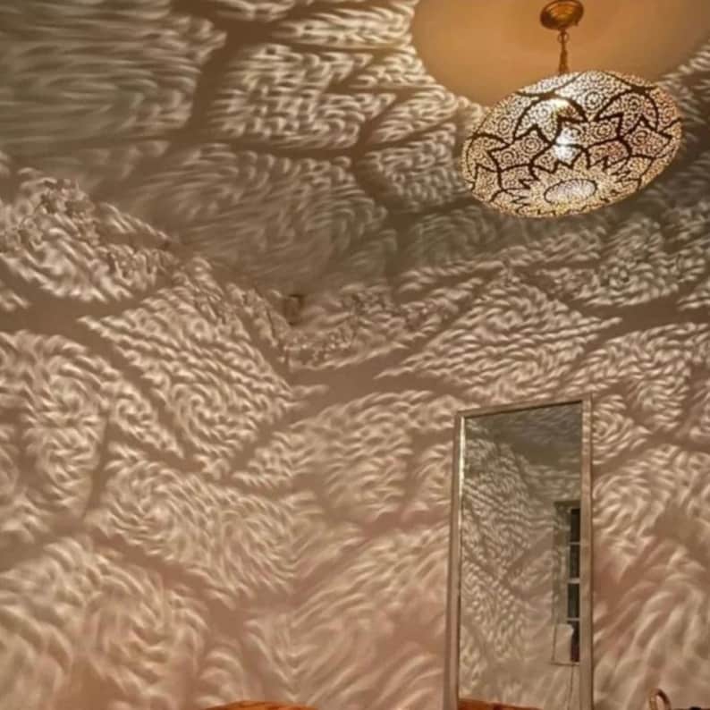 May include: An ornate, gold-colored pendant light fixture with intricate cut-out patterns hangs from the ceiling. The light casts a textured pattern on the walls, creating a unique visual effect. A mirror is visible below.
