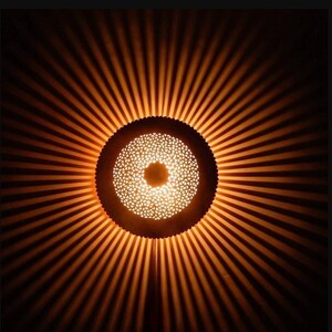 Op de afbeelding: Een ronde, decoratieve lamp met een geometrisch patroon. De lamp is gemaakt van metaal en heeft een warme, gouden gloed. De lamp werpt een lichtpatroon op de muur erachter.
