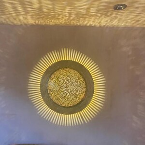 Op de afbeelding: Een ronde, gouden plafondlamp met een decoratief, gepatroneerd ontwerp. Het licht werpt een warme gloed op het omringende plafond.
