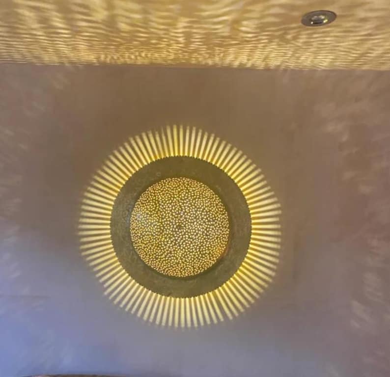 Op de afbeelding: Een ronde, gouden plafondlamp met een decoratief, gepatroneerd ontwerp. Het licht werpt een warme gloed op het omringende plafond.