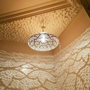 May include: An ornate, oval-shaped pendant light with a floral pattern, casting intricate shadows on the wall. The light fixture is suspended from the ceiling, illuminating the room with a warm glow. The ceiling and walls are painted in a neutral color.
