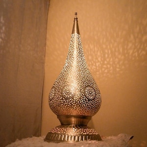 K&ouml;nnte beinhalten: Eine metallische, tropfenf&ouml;rmige Tischleuchte mit einem komplizierten, lasergeschnittenen geometrischen Muster. Die Lampe verf&uuml;gt &uuml;ber einen spitzen Abschluss an der Oberseite und einen breiten, runden Sockel mit vertikalen Rillen.
