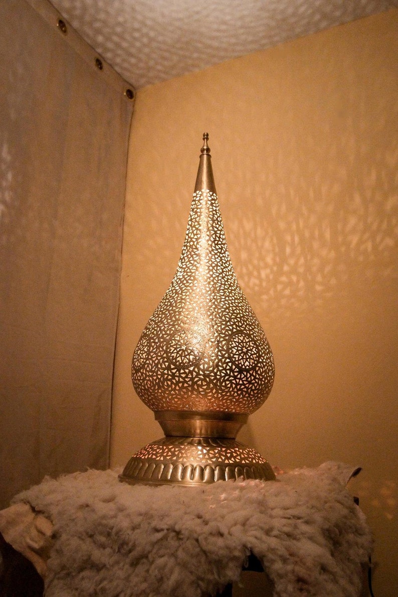K&ouml;nnte beinhalten: Eine metallische, tropfenf&ouml;rmige Tischleuchte mit einem komplizierten, lasergeschnittenen geometrischen Muster. Die Lampe verf&uuml;gt &uuml;ber einen spitzen Abschluss an der Oberseite und einen breiten, runden Sockel mit vertikalen Rillen.