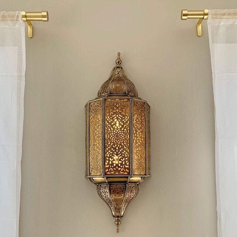 Może przedstawiać: Złota metalowa lampa ścienna z misternymi wzorami wyciętymi. Lampa ma tradycyjny marokański styl i jest oświetlona ciepłym białym światłem.