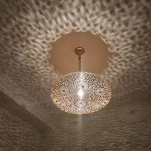 May include: A round, gold-colored pendant light fixture with a decorative, intricate pattern. The light fixture is hanging from a ceiling and casting a patterned shadow on the wall.