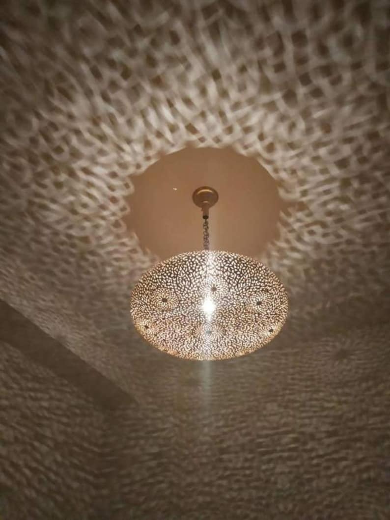 May include: A round, gold-colored pendant light fixture with a decorative, intricate pattern. The light fixture is hanging from a ceiling and casting a patterned shadow on the wall.