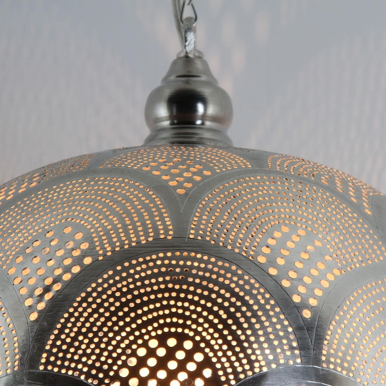 May include: A silver-toned metal pendant light fixture with a dome shape. The surface features an intricate pattern of small, circular perforations arranged in repeating fan-like arcs, allowing light to shine through the metallic material.
