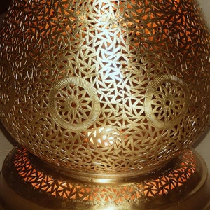 K&ouml;nnte beinhalten: Eine goldfarbene Metalllampe mit abgerundeter, bauchiger Form, die mit komplizierten, lasergeschnittenen geometrischen Mustern und kreisf&ouml;rmigen Blumenmotiven versehen ist. Die Oberfl&auml;che ist perforiert und l&auml;sst warmes Licht durch.