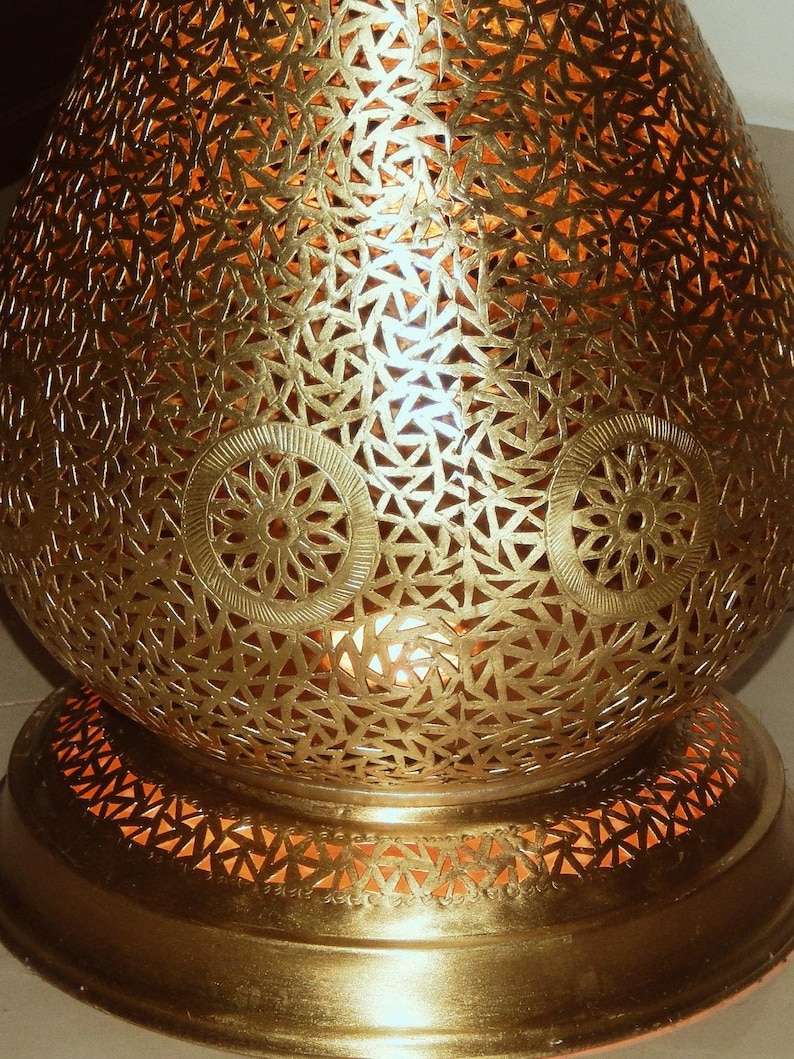 K&ouml;nnte beinhalten: Eine goldfarbene Metalllampe mit abgerundeter, bauchiger Form, die mit komplizierten, lasergeschnittenen geometrischen Mustern und kreisf&ouml;rmigen Blumenmotiven versehen ist. Die Oberfl&auml;che ist perforiert und l&auml;sst warmes Licht durch.