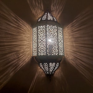 Może przedstawiać: Biała marokańska lampa wisząca z metalu z misternymi wycięciami. Lampa jest zapalona i emituje ciepłe światło. Tekst "NOURLamps" jest widoczny w prawym dolnym rogu obrazu.