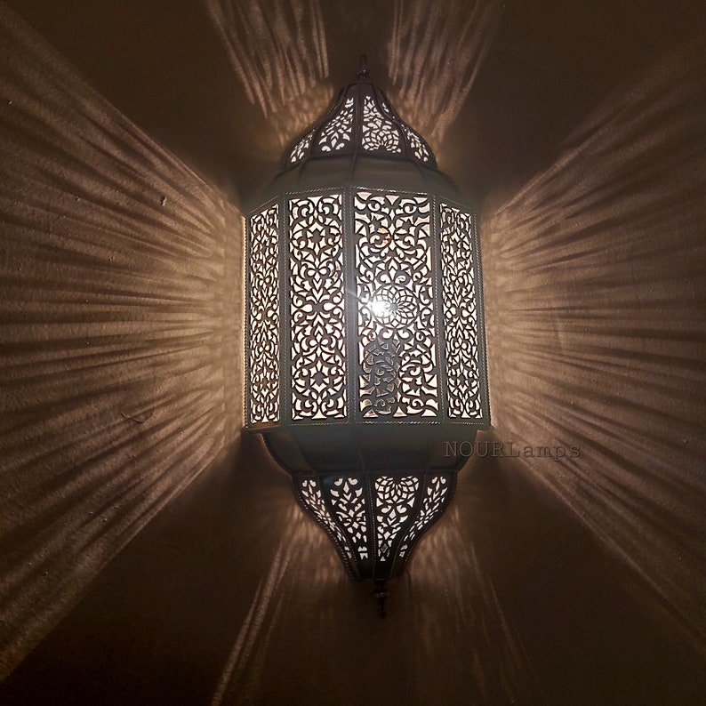 Może przedstawiać: Biała marokańska lampa wisząca z metalu z misternymi wycięciami. Lampa jest zapalona i emituje ciepłe światło. Tekst "NOURLamps" jest widoczny w prawym dolnym rogu obrazu.