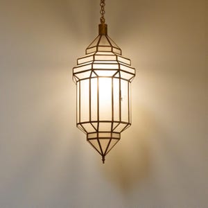Puede incluir: Una lámpara colgante con diseño geométrico. Tiene un marco de color latón y paneles de vidrio esmerilado. La lámpara emite una luz cálida, adecuada para la iluminación interior. El diseño recuerda a una linterna.