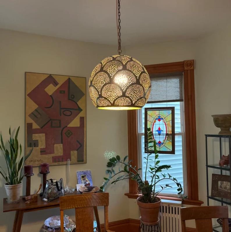 May include: A dome-shaped pendant light fixture with a dark exterior featuring a repeating scalloped pattern of small, circular perforations. The light is suspended by a metal chain and emits a warm, glowing light through the cutouts.