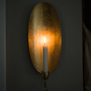 May include: A gold metal wall sconce with an oval-shaped backplate and a candle holder. A white candle is lit inside the sconce, casting a warm glow.
