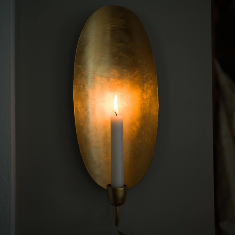 May include: A gold metal wall sconce with an oval-shaped backplate and a candle holder. A white candle is lit inside the sconce, casting a warm glow.