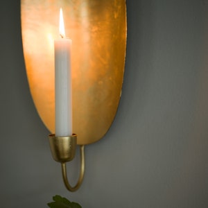 May include: A gold metal wall sconce with a candle holder and a lit white candle. The sconce has a unique oval shape and a smooth, polished finish.