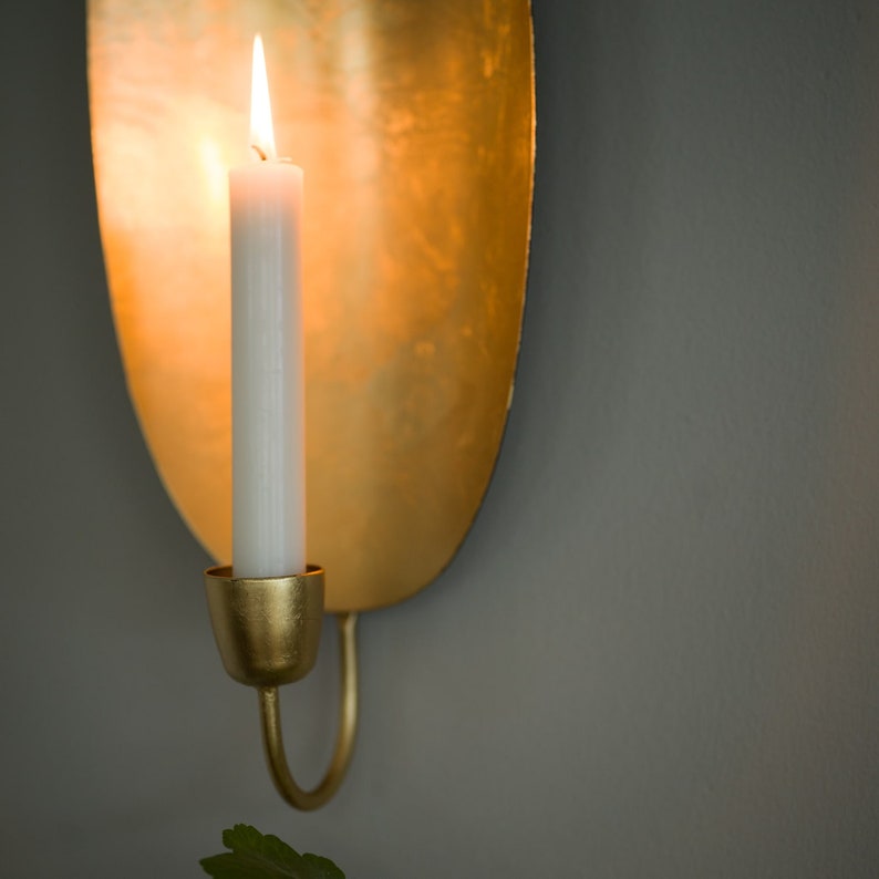 May include: A gold metal wall sconce with a candle holder and a lit white candle. The sconce has a unique oval shape and a smooth, polished finish.