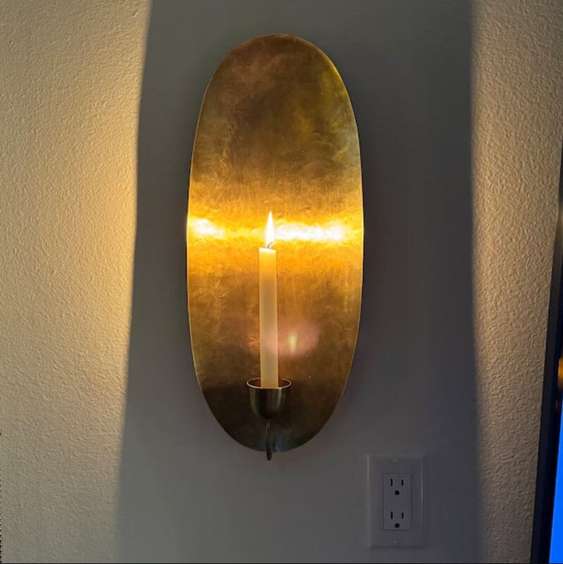 May include: A gold oval wall sconce with a lit candle inside. The sconce is mounted on a white wall.
