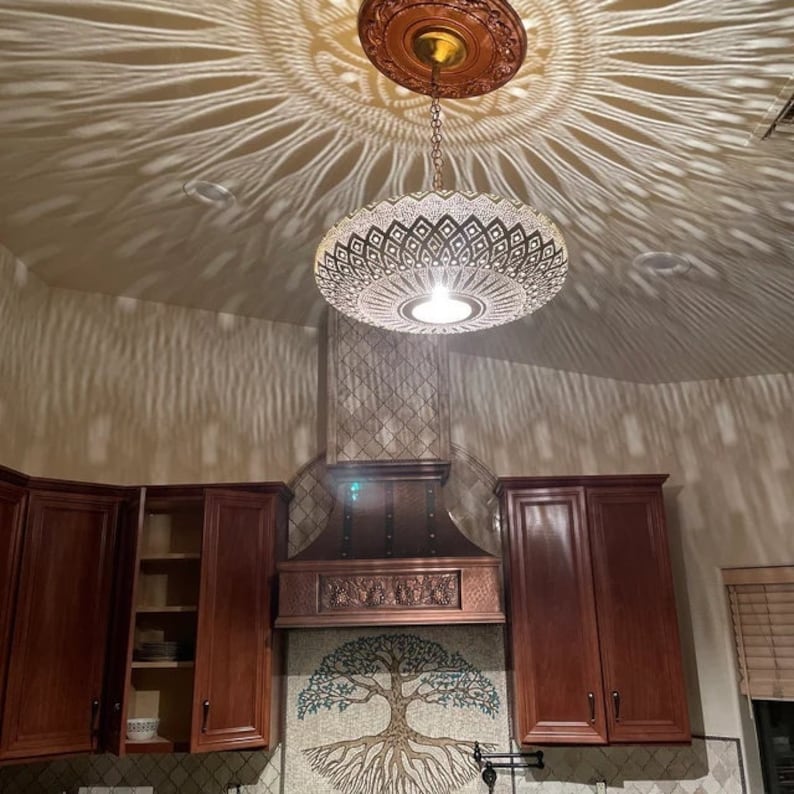 Puede incluir: Una gran l&aacute;mpara de techo de metal dorado adornada con un patr&oacute;n geom&eacute;trico cuelga de una cadena en una cocina. La l&aacute;mpara proyecta una sombra decorativa en el techo. La cocina tiene gabinetes marrones y una campana de cocina de cobre con un salpicadero de mosaico que presenta un dise&ntilde;o de &aacute;rbol de la vida.