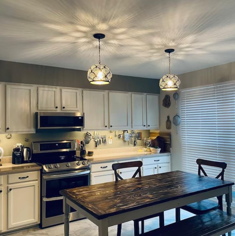 May include: Two dome-shaped pendant lights with a metallic, scalloped pattern hang from the ceiling, casting intricate light patterns. The kitchen features white cabinets, a stainless steel stove, and a dark wood dining table with matching benches.