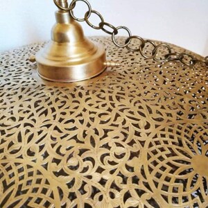 May include: A brass pendant light fixture with a decorative, intricate, cut-out design. The fixture has a chain for hanging and a brass cap.