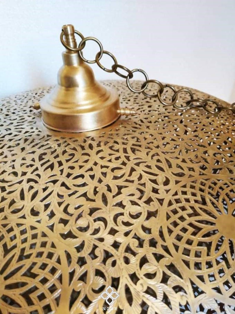 May include: A brass pendant light fixture with a decorative, intricate, cut-out design. The fixture has a chain for hanging and a brass cap.