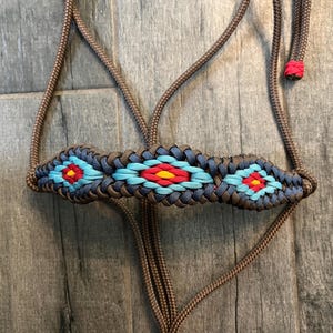 May include: A brown paracord halter featuring a noseband with a woven geometric pattern in dark blue, light blue, red, and yellow. The halter is constructed from durable cord with a matching brown lead and a small red accent at the end of the strap.