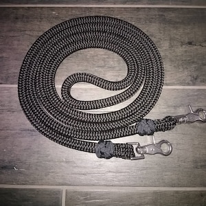 May include: Black braided rope reins with silver metal snap hooks. The reins are coiled and laid out on a wood surface.