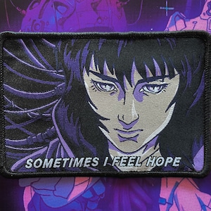 Könnte beinhalten: Ein schwarzer und lilafarbener gestickter Aufnäher mit einer Nahaufnahme des Gesichts einer Frau. Die Frau hat dunkles Haar und blickt direkt auf den Betrachter. Der Text "SOMETIMES I FEEL HOPE" ist unter dem Bild gedruckt.