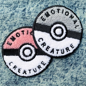 May include: Two embroidered patches in the shape of a Pokeball. The top patch is gray with the words "Emotional Creature" in black. The bottom patch is pink with the words "Emotional Creature" in black.