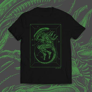 May include: Black t-shirt with a green illustration of an Alien creature inside a rectangular frame. The design includes anatomical labels and the word "ALIEN". The background features a matching green alien design.