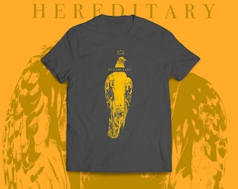 May include: Dark gray t-shirt with a yellow graphic of a bird wearing a crown. The word "HEREDITARY" is printed above the shirt, and the word "HEREDITARY" is printed below the crown. The background is yellow with a bird wing design.