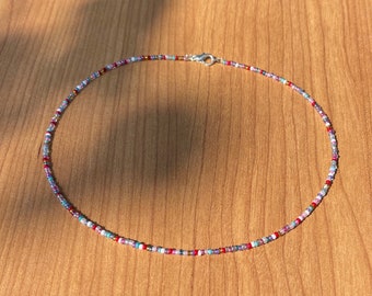 May include: A delicate beaded necklace with a variety of small, colorful beads. The beads are in shades of red, blue, purple, and white, strung together on a thin cord. The necklace has a silver clasp and is displayed on a wooden surface.