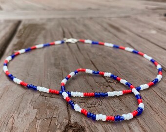 May include: A beaded necklace and bracelet set. The jewelry features a pattern of red, white, and blue beads. The necklace has a silver clasp. The jewelry is displayed on a weathered wooden surface.