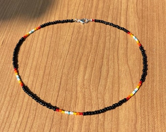 May include: A beaded necklace featuring a black base with a colorful pattern of red, orange, yellow, and turquoise beads. The necklace has a silver clasp and is displayed on a wooden surface.