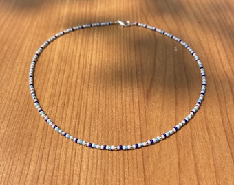 May include: A beaded necklace with a silver clasp. The necklace features small, round beads in shades of blue, white, and silver. The necklace is displayed on a wooden surface.