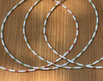 May include: Four delicate beaded necklaces with white, gold, turquoise, and pink beads. Each necklace has a silver clasp. The necklaces are displayed on a wooden surface.