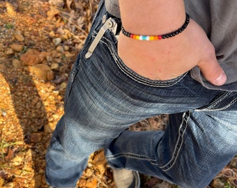 May include: A black beaded bracelet with a colorful stripe of red, orange, yellow, turquoise, and white beads. The bracelet is worn on a wrist, with blue jeans and a gray shirt in the background.