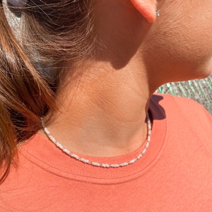 May include: A delicate beaded necklace with white and gold-colored beads. The necklace is worn over a coral-colored t-shirt. A small, square earring is visible in the ear.