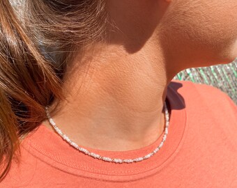 May include: A delicate beaded necklace with white and gold-colored beads. The necklace is worn over a coral-colored t-shirt. A small, square earring is visible in the ear.