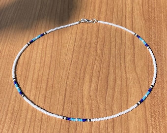 May include: A beaded necklace with a white base, accented with blue and black beads. The necklace has a silver clasp and is displayed on a wooden surface. The beads are small and evenly spaced, creating a delicate, minimalist design.